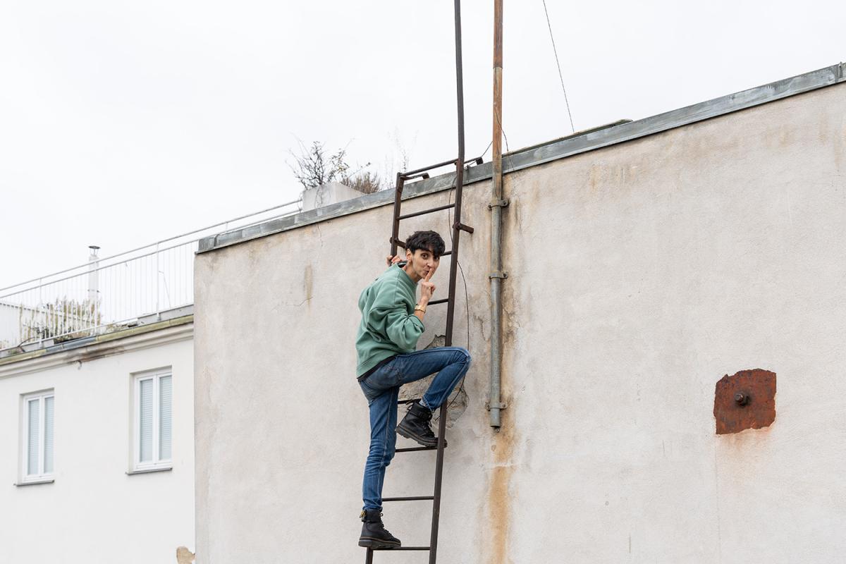 A person climbs a metal ladder on a building wall, smiling over their shoulder while placing a finger to their lips.