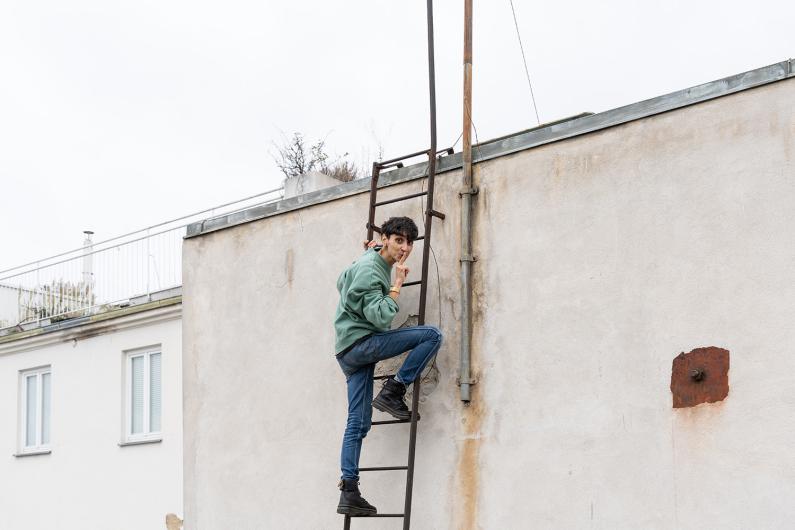 A person climbs a metal ladder on a building wall, smiling over their shoulder while placing a finger to their lips.
