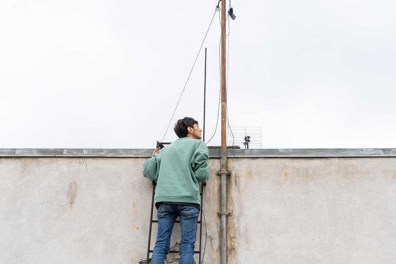 A person climbs a metal ladder on a building wall, looking up intently at an antenna pole above.