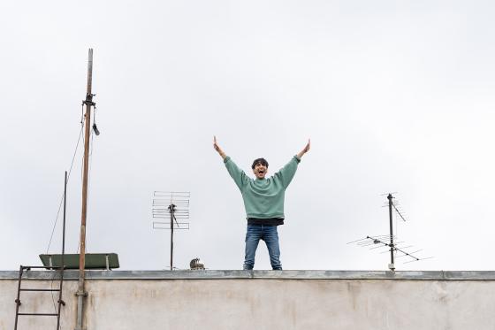 A person stands on a rooftop between several antennas, joyfully raising both arms in the air under a gray open sky.