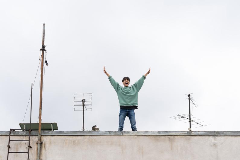 A person stands on a rooftop between several antennas, joyfully raising both arms in the air under a gray open sky.