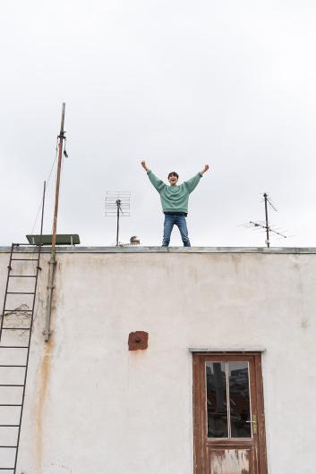 A person stands on a rooftop with both arms raised and a wide smile, surrounded by old antennas and a metal ladder.