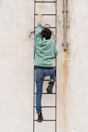 Eine Person in Jeans und grünem Pullover klettert eine Metallleiter hinauf, die an einer hellen Hauswand befestigt ist. Der Fokus liegt auf der Bewegung nach oben.