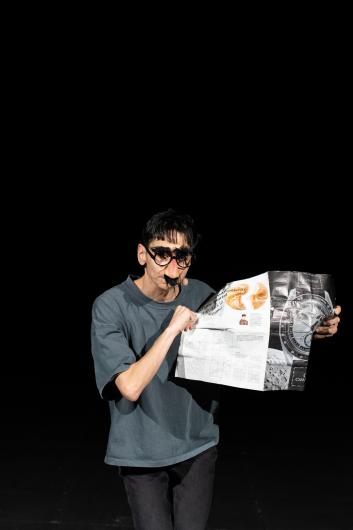 The person stands facing forward holding an unfolded newspaper. They wear a conspicuous pair of glasses with an attached nose. Their gaze is directed downward at the newspaper while the background is completely black.