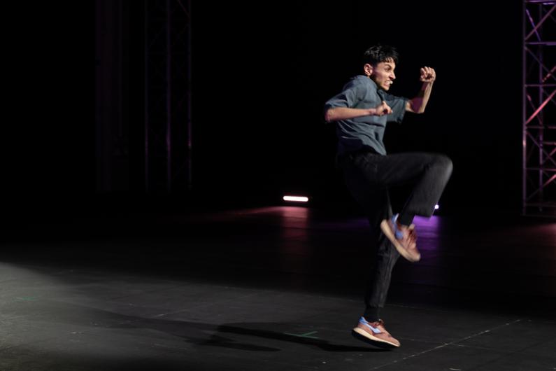 The person is captured in a powerful jump across the stage. One knee is lifted and the arms are bent. Colored stage lighting and a metal truss are visible in the background.