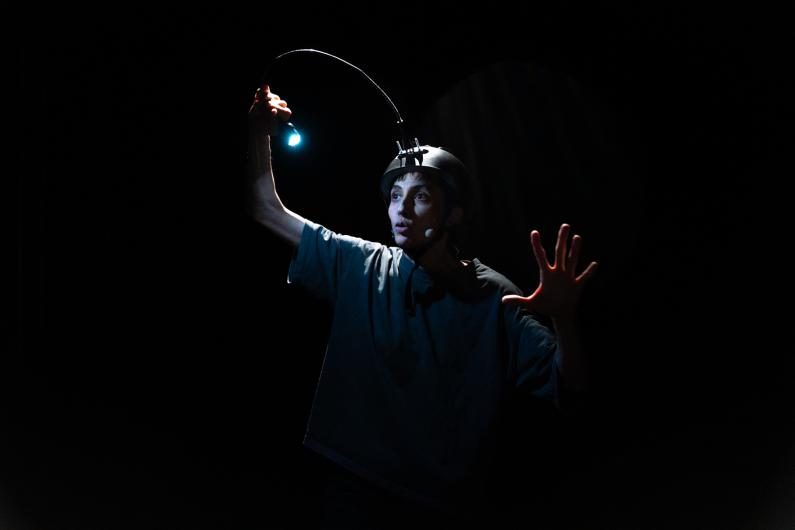 A performing person stands in darkness wearing a helmet with a flexible rod ending in a small light. One hand lifts the light upward while the other hand is open and extended forward. The face is clearly illuminated while the background remains black.