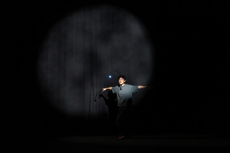 A single person stands with arms outstretched inside a large oval cone of light on a dark stage. A small light glows above the helmet. A curtain is faintly visible in the background.