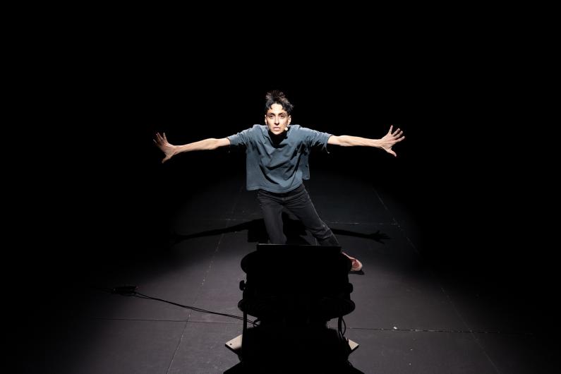 The person faces forward on stage with both arms spread wide. The body is lit from above while the floor and background remain dark. A technical object appears faintly in the foreground.