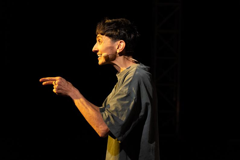 A performing person is shown in side profile wearing a headset microphone. Two fingers point forward. The face is warmly lit while the background stays black.