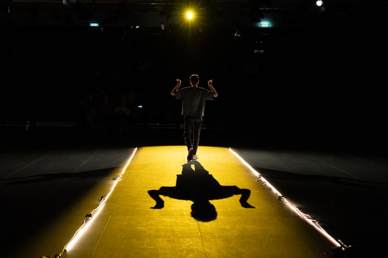 Die Person geht von hinten über einen schmalen, gelb beleuchteten Laufsteg in einen dunklen Zuschauerraum. Beide Arme sind angewinkelt und angehoben. Ein starkes Bühnenlicht wirft einen deutlichen Schatten auf den Boden.