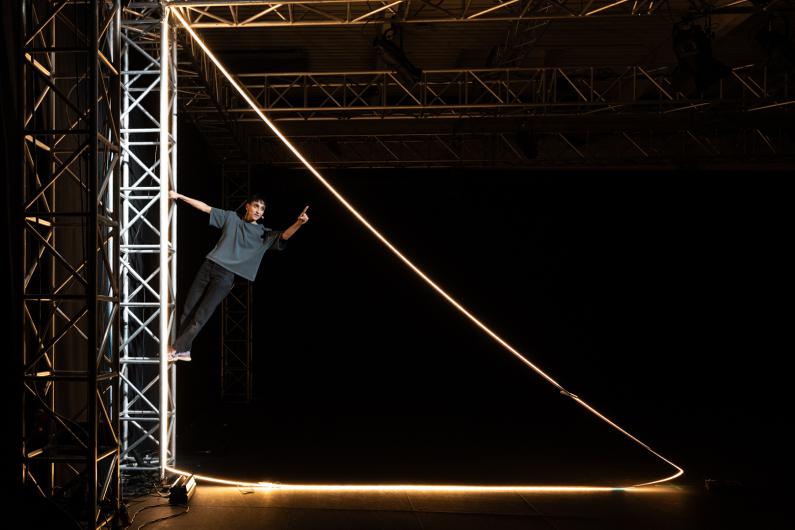 Die Person hält sich seitlich an einem hohen Metallgerüst fest und lehnt sich in den Raum. Eine helle Lichtlinie verläuft diagonal vom Gerüst über den Bühnenboden und rahmt die Szene geometrisch ein.