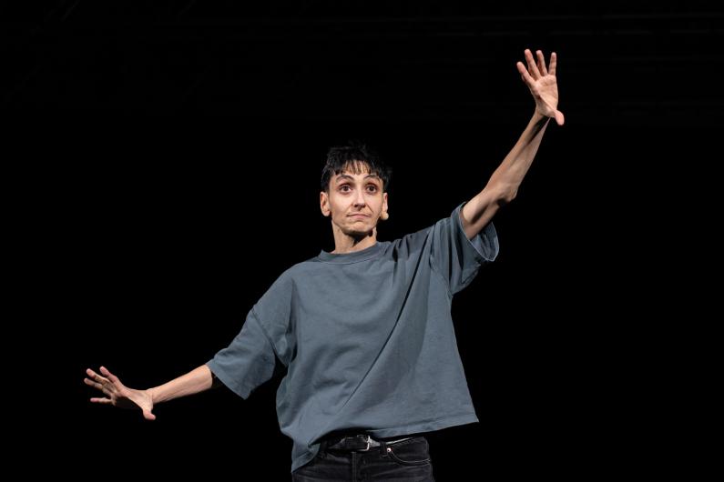 The person stands on stage with arms slightly raised and hands open. The body is clearly lit while the background remains completely dark.