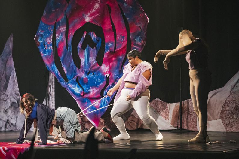 Drei Performer*innen agieren auf der Bühne vor einer großen, farbintensiven Kulisse. Eine Person kriecht am Boden, eine andere zieht sie mit elastischen Bändern, während eine dritte sich eine Strumpfhose über den Kopf zieht.