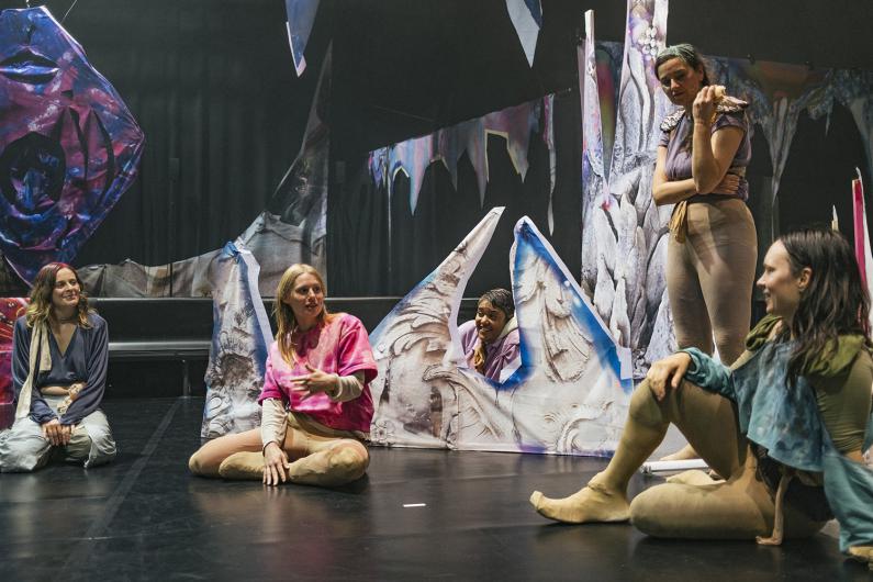 Five performers sit and stand within a stage set made of painted, jagged shapes. They talk and laugh together, creating a playful and communal atmosphere.