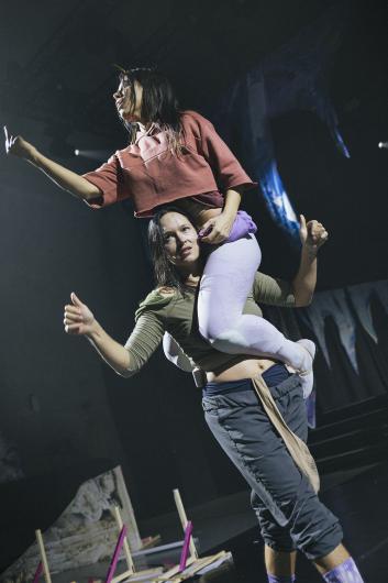 One performer carries another on their shoulders, both in motion with raised arms and expressive gestures. Dramatic light falls from above.