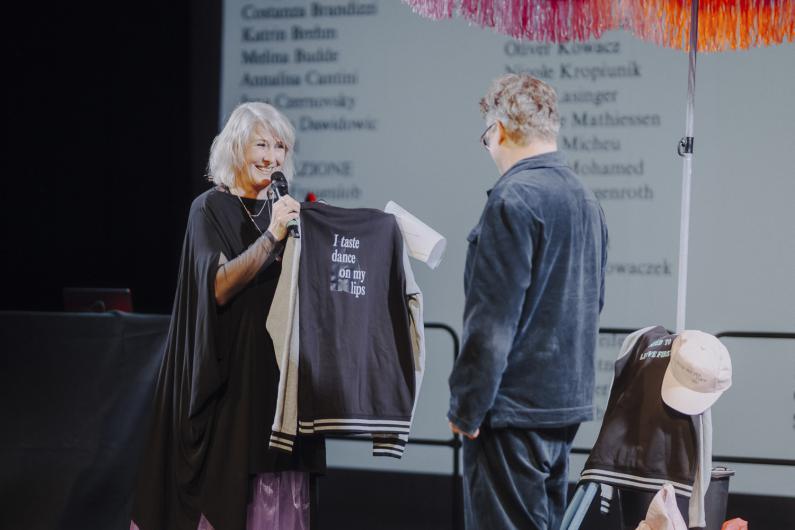 A person smiles while holding up a printed garment and presenting it to another person standing opposite; additional props are visible in the background.