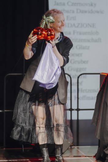 A person holds two shiny, cherry-shaped objects in their hands and smiles while turning slightly to the side; the stage lighting highlights the colours.