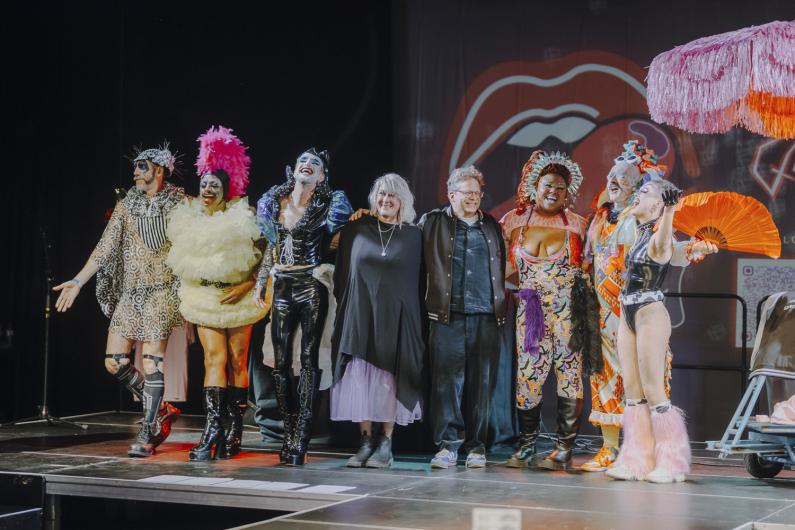 A group of performers in colourful and extravagant costumes stands on stage together with two others, facing the audience.