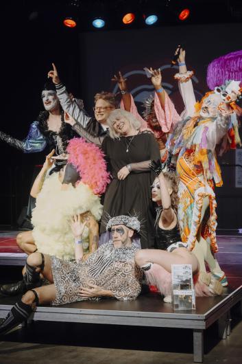 Several performers and participants pose together on stage, some raising their arms or gesturing while standing close together.