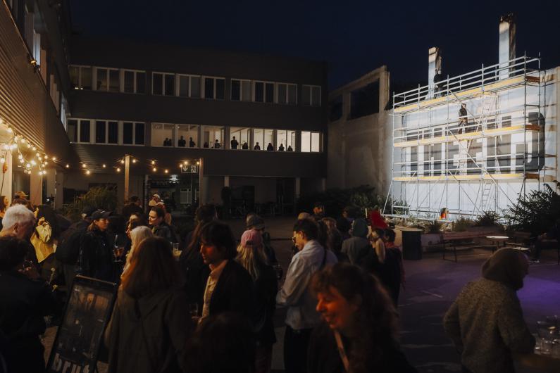 Ein nächtlicher Außenbereich mit vielen Besucher*innen, warmen Lichterketten und einer hell beleuchteten Gerüstkonstruktion, die als Bühne oder Installation dient.