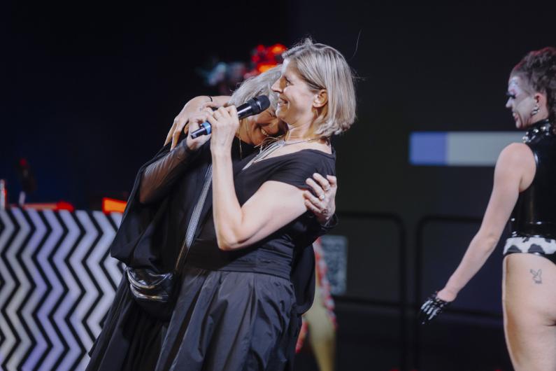 Two people embrace closely on stage, one holding a microphone, while other performers are visible in the background.