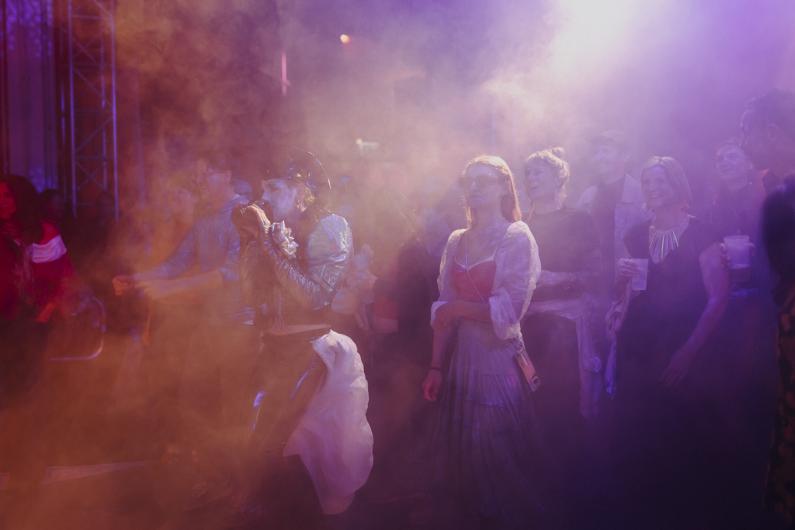 A dense crowd stands in front of the stage in coloured light and haze, while a performing person with a microphone is visible in the foreground.