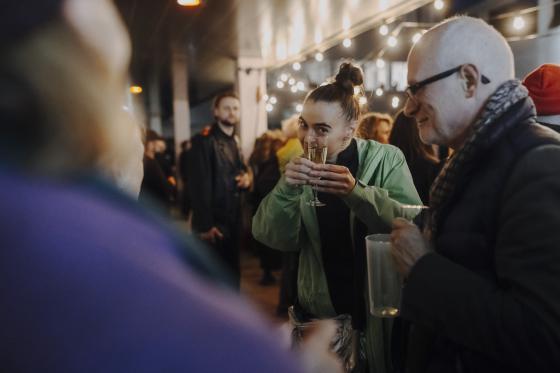 Two people in the foreground clink drinks while a dense, sociable crowd mingles in the background.