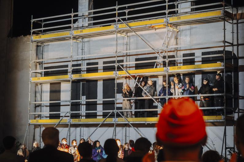 Several performers stand on a scaffold structure, looking down at the audience gathered below.
