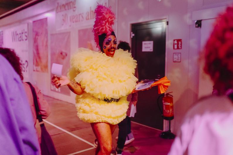 A drag performer in a striking yellow feathered costume moves through a pink-lit indoor space, engaging with the audience.