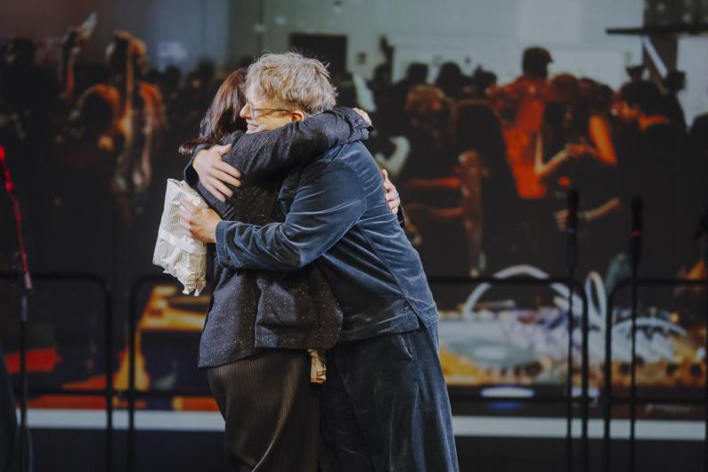Two people stand on stage embracing in front of a large projection showing a crowd.