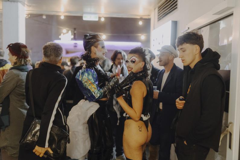 Mehrere Personen in aufwendigen Drag-Outfits und mit starkem Bühnen-Make-up stehen dicht beieinander in einem Innenraum, lachen und unterhalten sich in lebhafter Atmosphäre.