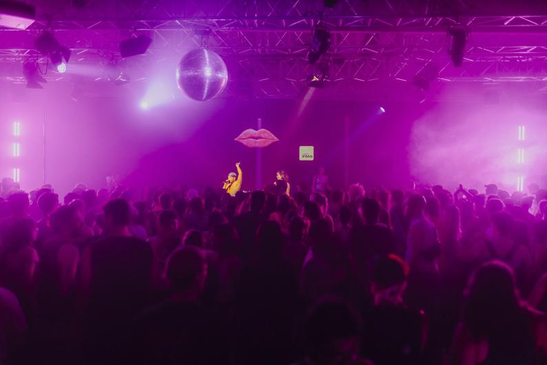 Wide shot of the venue with a large crowd. The stage with a glowing lips motif is centrally visible, with pink light filling the space.