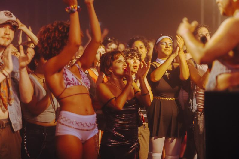 Audience stands closely in front of the stage and applauds. Faces are visible in warm light, many looking attentively forward.