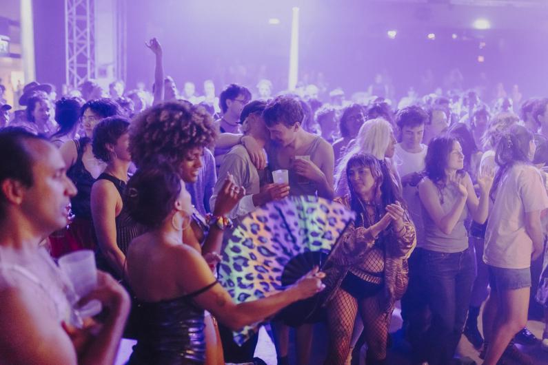 Tanzende Besucher*innen füllen den Raum unter pink-violettem Licht. Im Vordergrund hält eine Person einen auffälligen Fächer, während andere klatschen und sich bewegen.