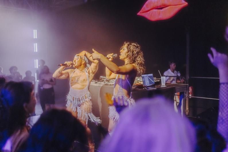 Two performers stand on stage with microphones, performing together. A lips motif glows in the background, with the audience visible in the foreground.