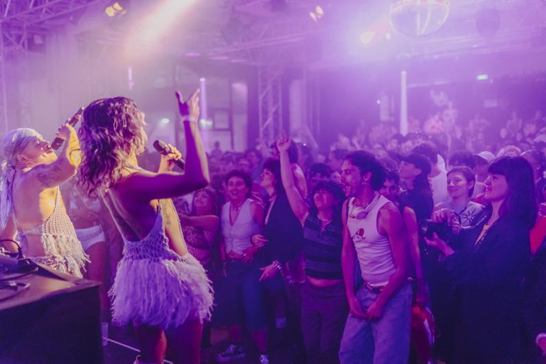 Blick von der Bühne ins Publikum: Zwei Performer*innen stehen vor einer großen, dicht gedrängten Menge. Scheinwerfer und Nebel erzeugen eine atmosphärische Lichtstimmung.