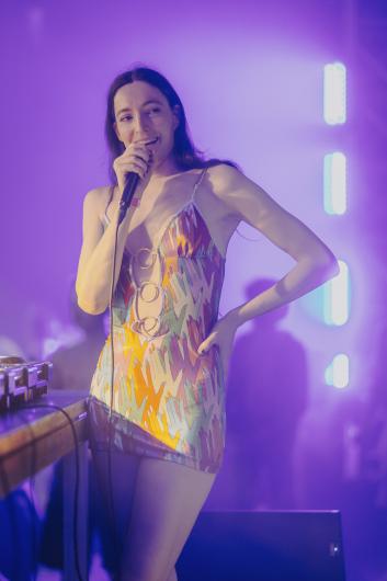 Person stands on stage holding a microphone, smiling toward the audience. They wear a short, colourful patterned dress. The scene is bathed in purple light, with light bars and a DJ booth visible in the background.