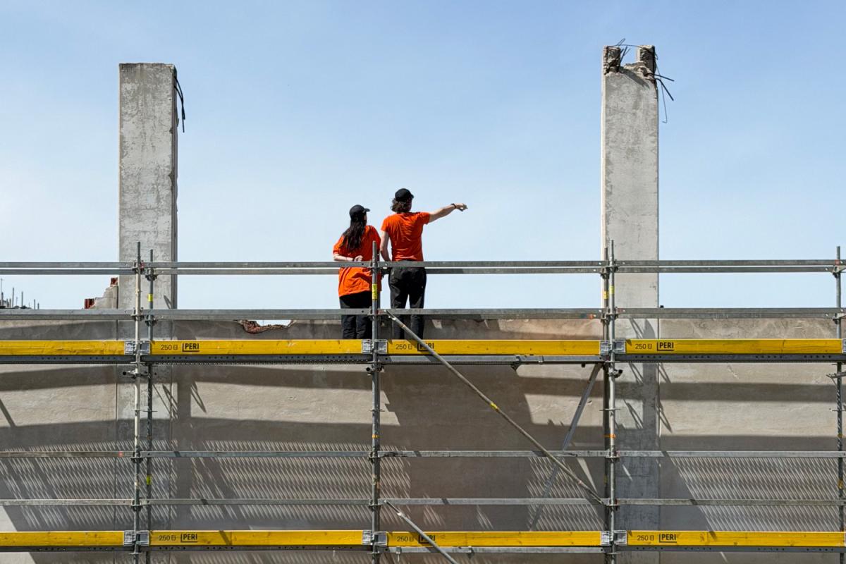Zwei Personen in orangefarbenen Oberteilen und dunklen Kappen stehen oben auf einem Baugerüst vor einer hellen Wand und blicken in die Ferne. Eine der beiden zeigt mit ausgestrecktem Arm nach rechts. Hinter ihnen ragen zwei hohe Betonpfeiler in den klaren blauen Himmel, während das Gerüst mit gelben Querbalken und silbernen Streben das Bild geometrisch strukturiert.
