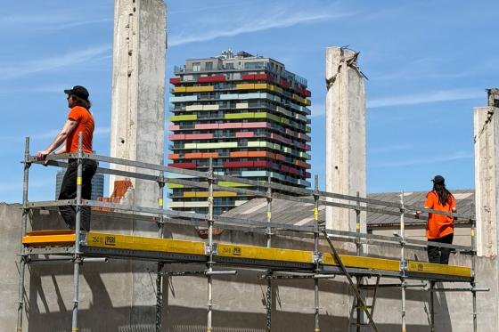 Zwei Personen in orangefarbenen T-Shirts und schwarzen Kappen stehen auf einem Baugerüst zwischen hohen, rohen Betonpfeilern. Im Hintergrund erhebt sich ein markantes Hochhaus mit bunten Balkonen in Rot, Gelb, Grün und Rosa vor einem klaren blauen Himmel. Die gelben Gerüstbalken und die industrielle Umgebung kontrastieren mit der farbigen Architektur im Hintergrund.