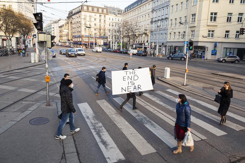 Several people are crossing a wide street junction on a zebra crossing. In the center, one person carries a large white sign with handwritten black letters reading “THE END IS NEAR.” Cars drive in the background, tram tracks cut through the street, and multi-storey buildings line the scene. Some people wear winter jackets and face masks.