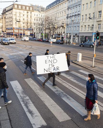 Nahaufnahme einer Straßenszene an einem Zebrastreifen. Im Vordergrund hält eine Person ein großes weißes Schild, auf dem in schwarzen Buchstaben „THE END IS NEAR“ steht, sodass der Oberkörper hinter dem Schild verborgen ist. Weitere Personen gehen über die Straße, eine Person in blauer Jacke trägt Einkaufssackerl. Im Hintergrund sind Häuserfassaden, Autos und Schienen zu sehen.