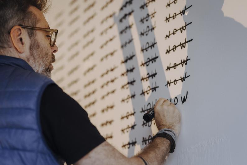 A person writes words on a white wall with a black marker while others are already crossed out. Light falls diagonally across the surface.