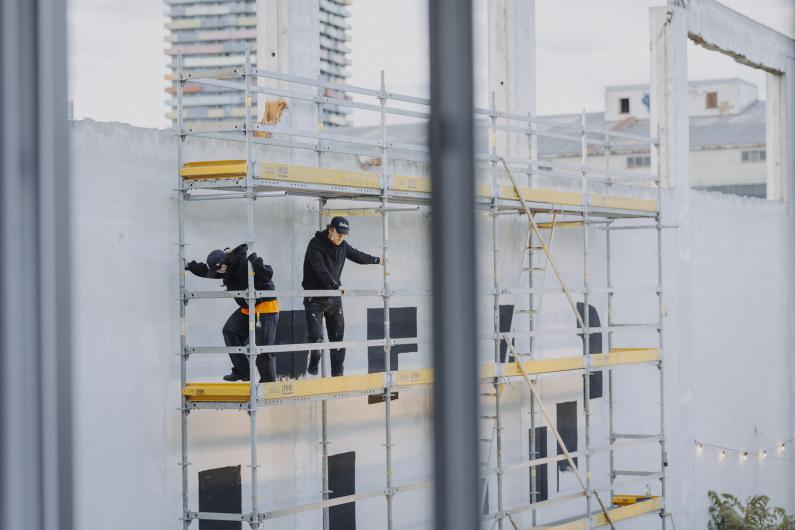 Two people move on scaffolding in front of a painted wall. The scene is partially obscured by a railing in the foreground.