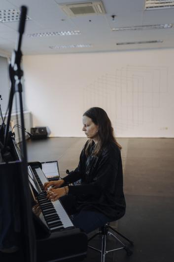 Eine Person sitzt am Klavier in einem Proberaum und spielt. Im Hintergrund ist eine Wand mit geometrischen Linienzeichnungen zu sehen.