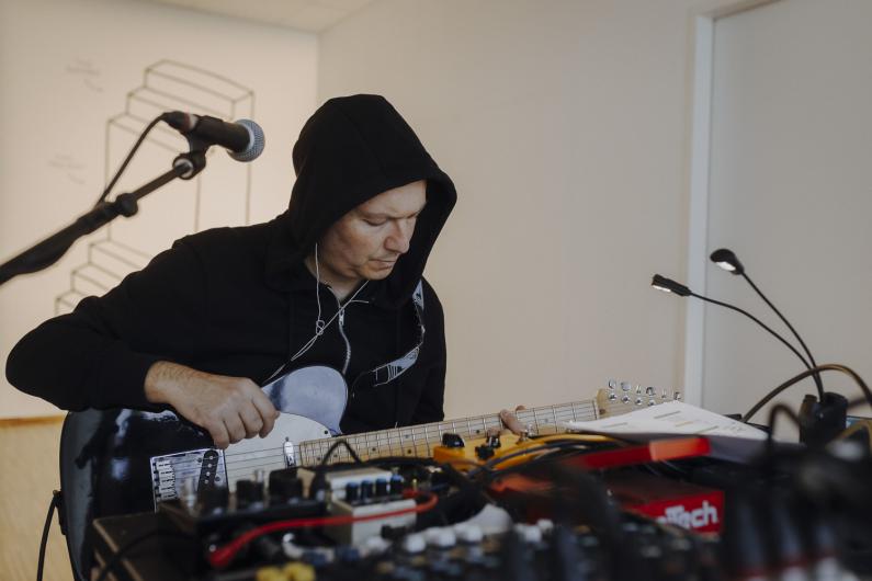 Eine Person mit Kapuzenpullover sitzt in einem Proberaum und spielt E-Gitarre. Vor ihr stehen Mikrofone und Effektgeräte, im Hintergrund ist eine Wand mit geometrischer Linienzeichnung zu sehen.