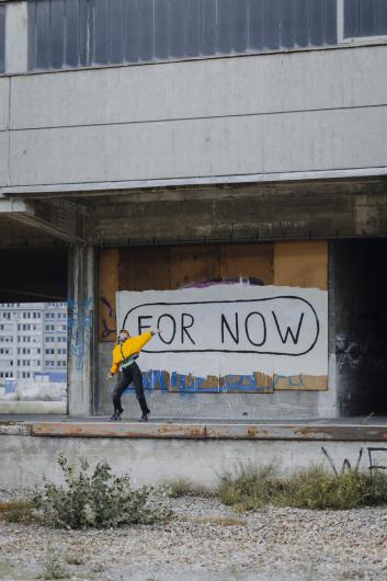 Eine Person in gelbem Pullover steht auf einer betonierten Plattform vor einer Wand mit dem Schriftzug „FOR NOW“. Die Umgebung wirkt industriell und leicht verwahrlost.