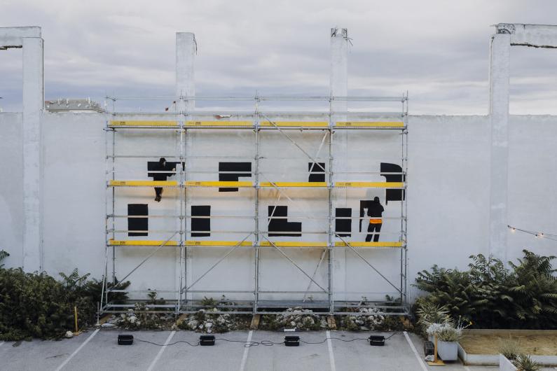 Two people stand on scaffolding in front of a large white wall, painting black geometric shapes. Plants and spotlights are placed on the ground below.