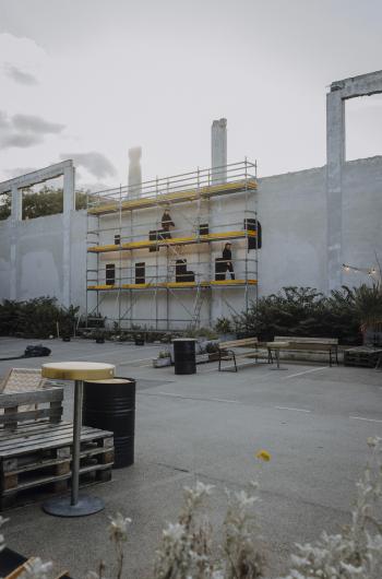 A view of the scaffolding in the brut courtyard where two people work on a wall piece. Tables and seating are visible in the foreground.