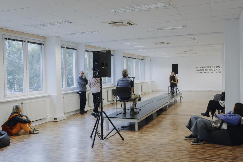 A bright room with a small stage: one person sits on a chair speaking into a microphone while several audience members are seated or standing around the space.