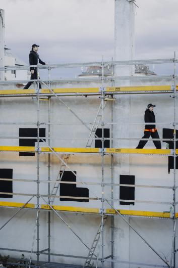 A man and a woman move along scaffolding in front of a wall with black geometric shapes. Both wear dark clothing and caps.